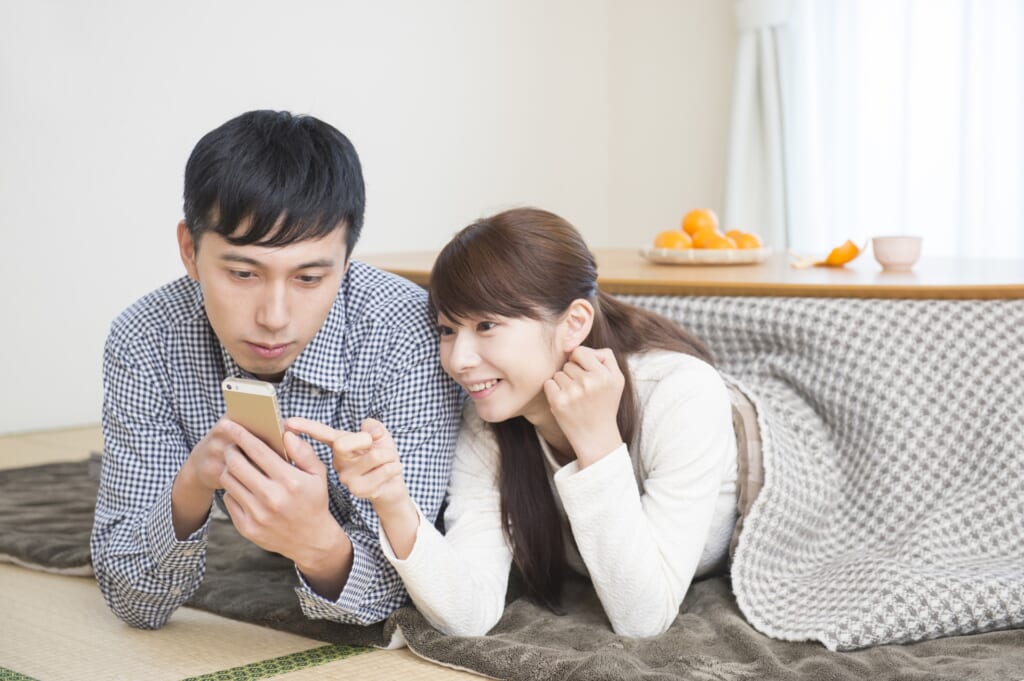 和室に設置されたこたつに入ってスマートフォンを見るカップル