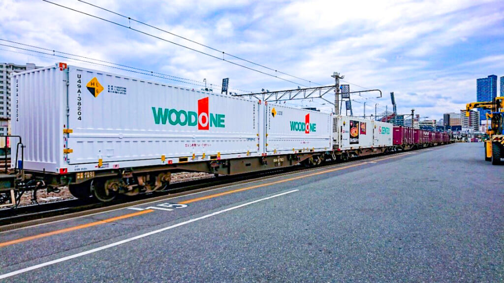 ウッドワンのロゴが入った白い専用コンテナを積んだ貨物列車が線路に沿って停車している様子。