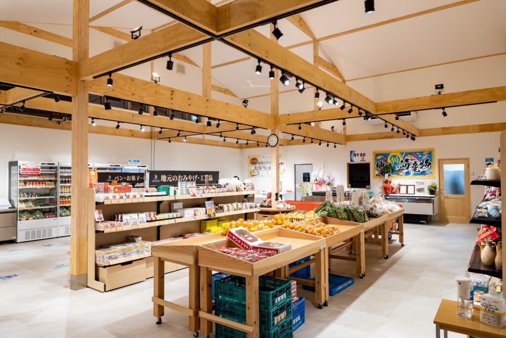 木材の梁がむき出しになった天井と明るい照明が特徴の、広々とした小売店内。中央には野菜や果物が並べられた木製の什器が複数あり、奥の棚には様々な加工品や冷蔵品が陳列されている。清潔感があり、自然素材を活かした温かい雰囲気の店舗空間。