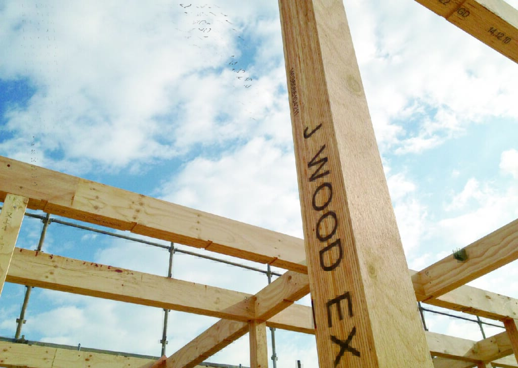 『J WOOD EX』と印字された木材の柱が中央に大きく映り、その周りに建設中の木造建築の骨組みが組まれている様子。晴れた日の屋外で、奥には青空が広がっている。