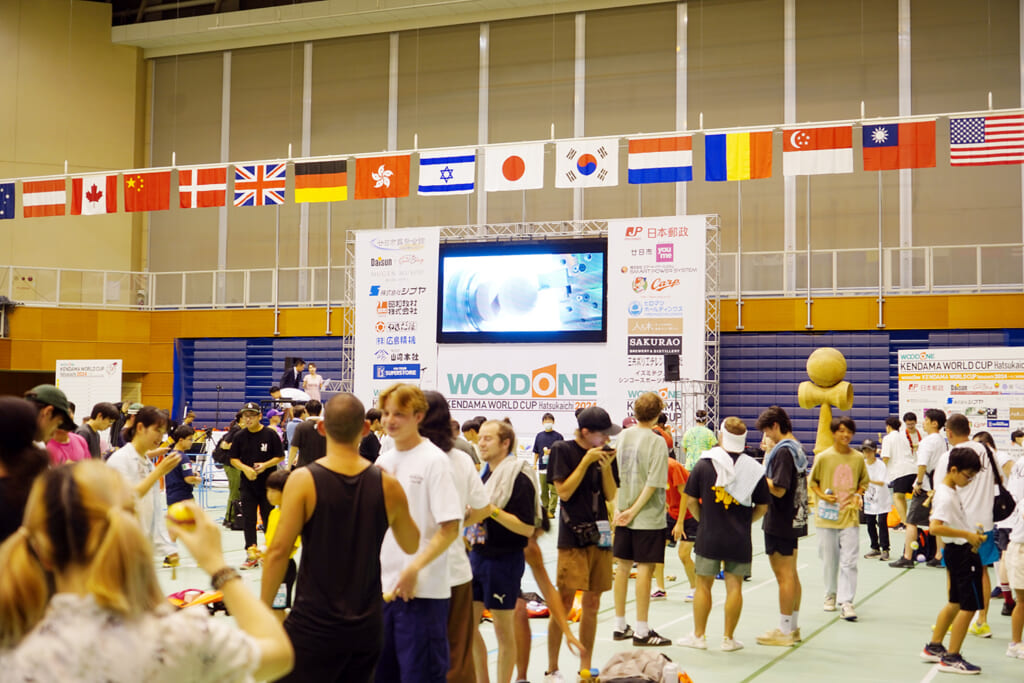 けん玉ワールドカップ廿日市 2024 の国際的な会場の様子。国旗の下で多くのプレイヤーが交流し、熱気にあふれている。