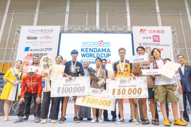 前回大会（2024年）ウッドワンけん玉ワールドカップ廿日市の授賞式。入賞者が賞金目録を持って笑顔で集合している様子。