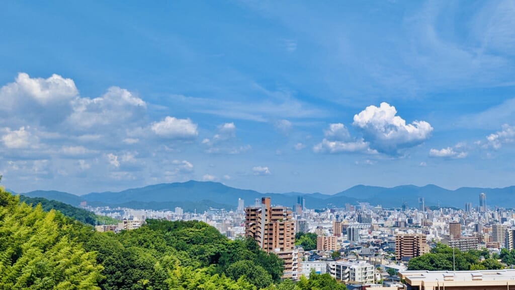 広島市の都市景観。緑の木々越しにマンションなどの建物群が広がり、遠景には山々が連なっている青空の風景写真。