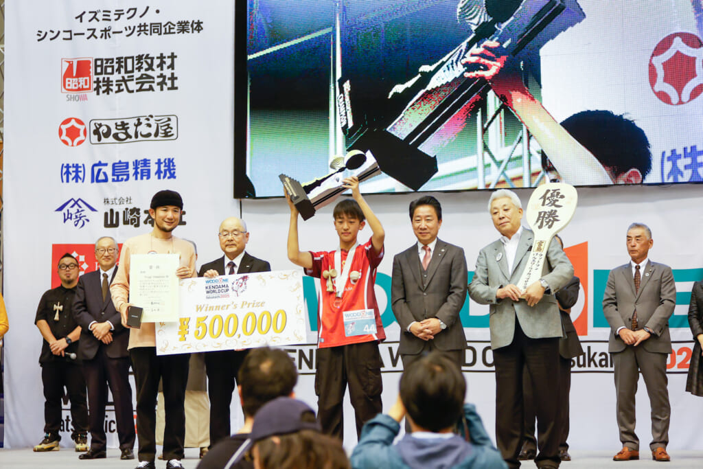 『ウッドワンけん玉ワールドカップ2025』表彰式の集合写真。大型モニターを背景に、優勝トロフィーや賞金ボードを持つ入賞者、大会関係者、そして公式マスコットキャラクターがステージに並び、大会の成功を祝っている様子。