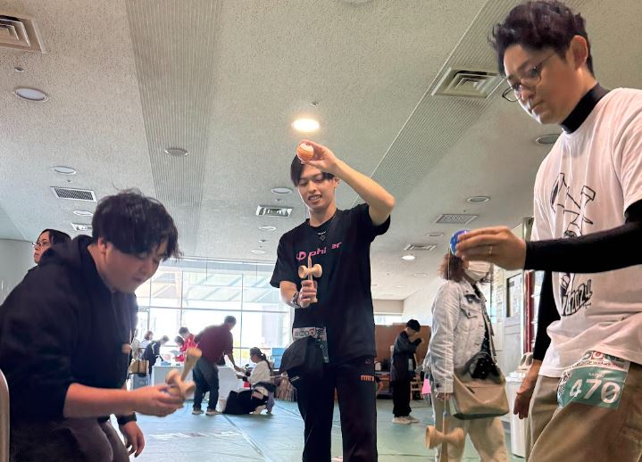 明るい屋内会場で、3人の男性がそれぞれ自分のけん玉を持ち、技の感触を確かめている。手前の選手は集中して玉を見つめ、背景では他の参加者たちが交流している、大会当日の熱気ある一幕。