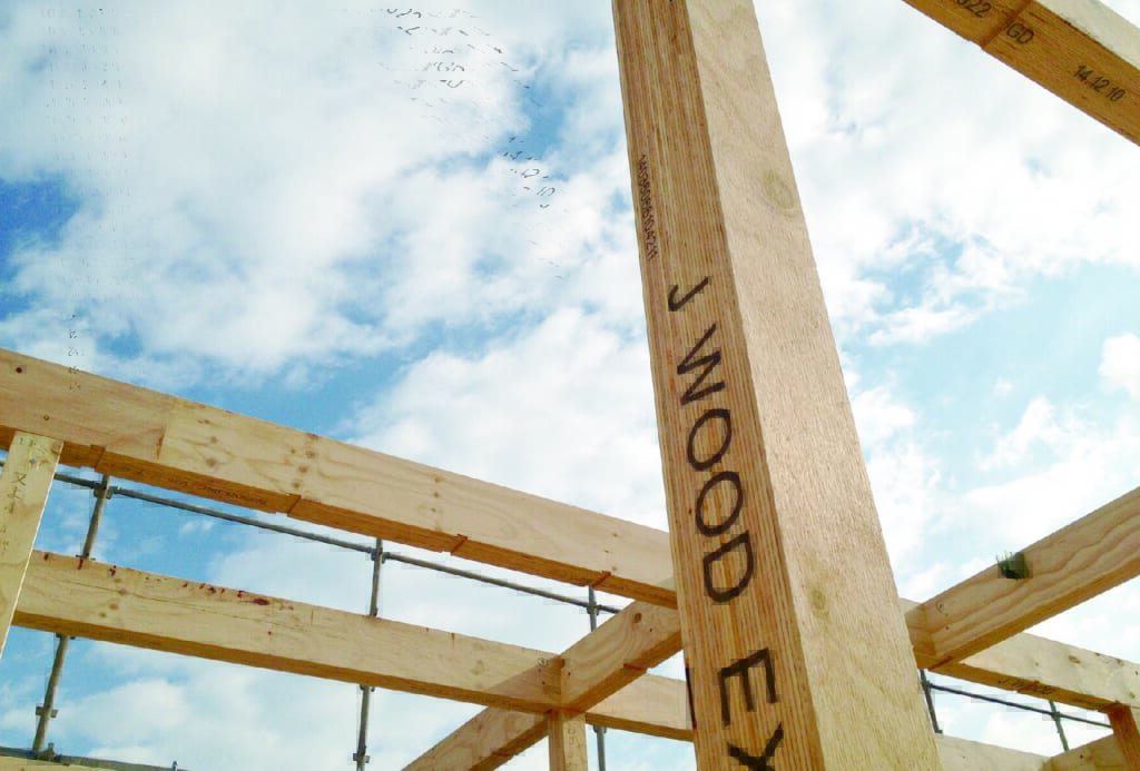 建設中の木造構造物。柱に『J WOOD EX』の文字が見え、明るい色の木材の骨組みが青い空に向かって組まれている。